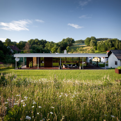 Streuobstwiesen schließen an den Garten an und spiegeln sich in der großflächigen Verglasung der Wohnräume – beinahe nahtlos gehen Wohnhaus, überdachte Terrasse, Rasen und Landschaft ineinander über