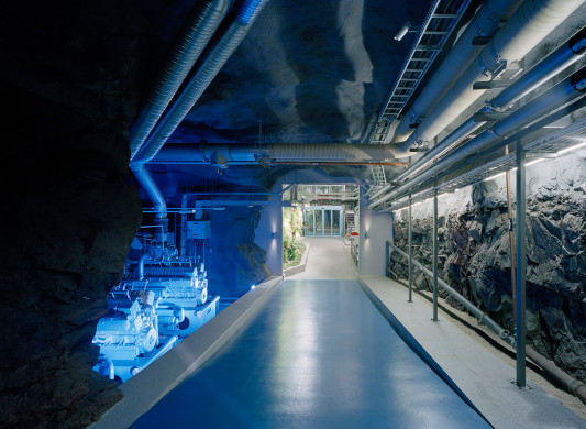 Ein Tunnel führt langsam in die Tiefe, flankiert von technischen Elementen wie einem Kühlturm, Rohrleitungen oder Dieselaggregaten