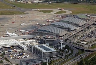 Aufsicht auf das Flughafengelände: Mittig ist das zylinderförmige, alte Parkhaus zu sehen, unter den drei Bogendächern befinden sich das Terminal 2, die Airport Plaza und das Terminal 1 (von links nach rechts)