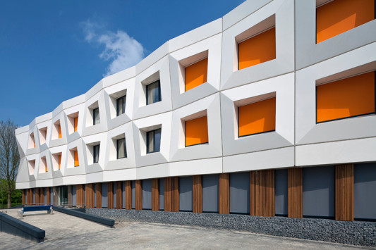 Die Fenster in der weißen, expressiv geformten Fassade des ersten und zweiten Obergeschosses sind mit Außenrollos in knalligem Orange ausgestattet