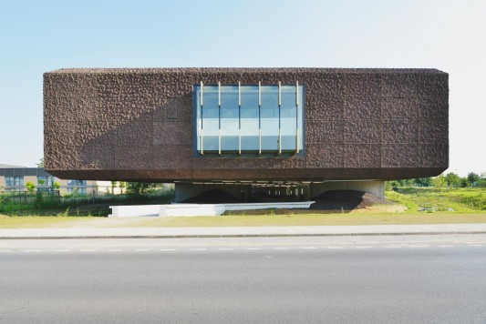 Die Fassade besteht aus dunklen, rotbraunen Betonelementen mit einem unregelmäßig gewellten Relief, durchbrochen von gläsernen Erkern