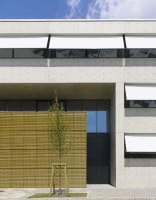 Die hellgraue Sichtbetonfassade wird von goldfarbenen Klappläden und weißen Markisen optisch aufgelockert