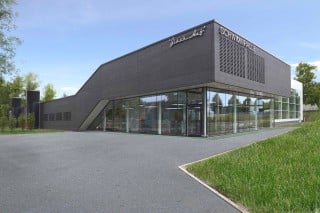 Im schwarzen Klinkerbau mit verglastem Sockel befinden sich der Eingang und das Foyer, dahinter folgen die Umkleiden, Duschen und das Lehrschwimmerbecken; der weiße Quader im Hintergrund nimmt das Schwimmerbecken auf (Nordostansicht)