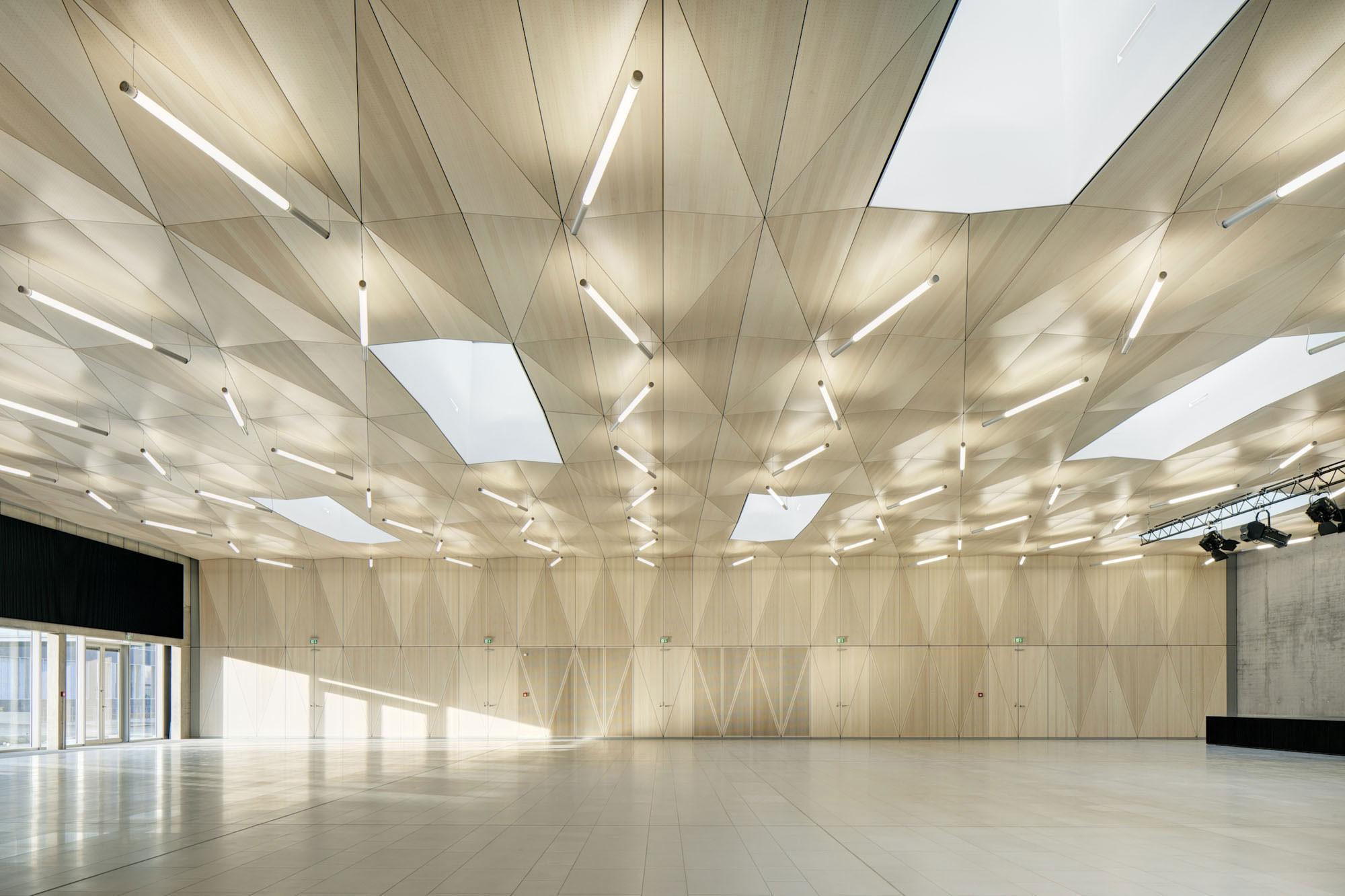 Die Decke und zwei Seiten der Aula sind aus überwiegend akustischen Gründen mit Holzpaneelen bekleidet, die ein rautenförmiges Faltwerk bilden und den riesigen, schachtelförmigen Raum zum Erlebnis werden lassen