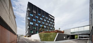 Der kompakte Neubau erhebt sich mit seinen sechs Geschossen auf einem teils eingegrabenen Sockel, rechts ist die kleine Betriebsstraße zu sehen, die von insgesamt vier Brücken überspannt wird (Südansicht)