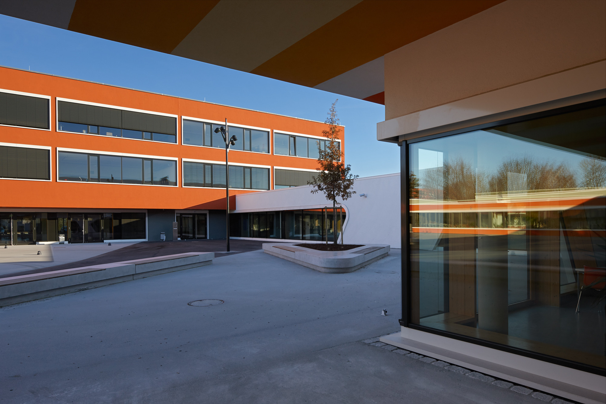 Der Pausenhof wird nun von allen Seiten eingefasst; der Bestand (hier das dreigeschossige Hauptgebäude) ist in kräftigen Farben verputzt, dazu kontrastieren die Neubauten mit ihren weißen Fassaden