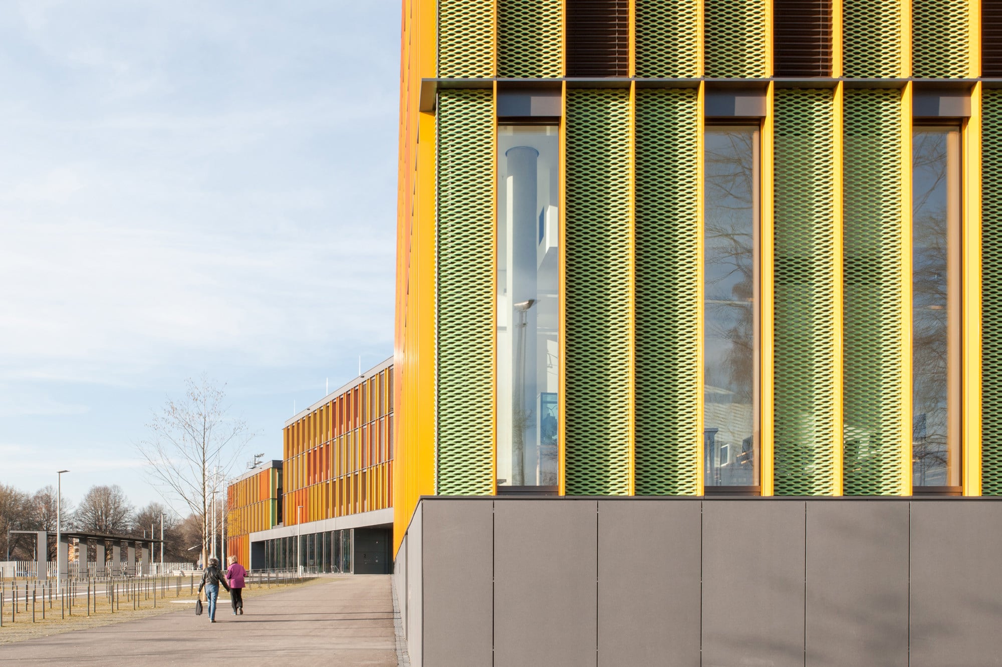 Orangefarbene Schwerter in engem Vertikalraster, dazwischen abwechselnd grüne Streckmetallelemente und dunkle Außenjalousien bestimmen die Fassade