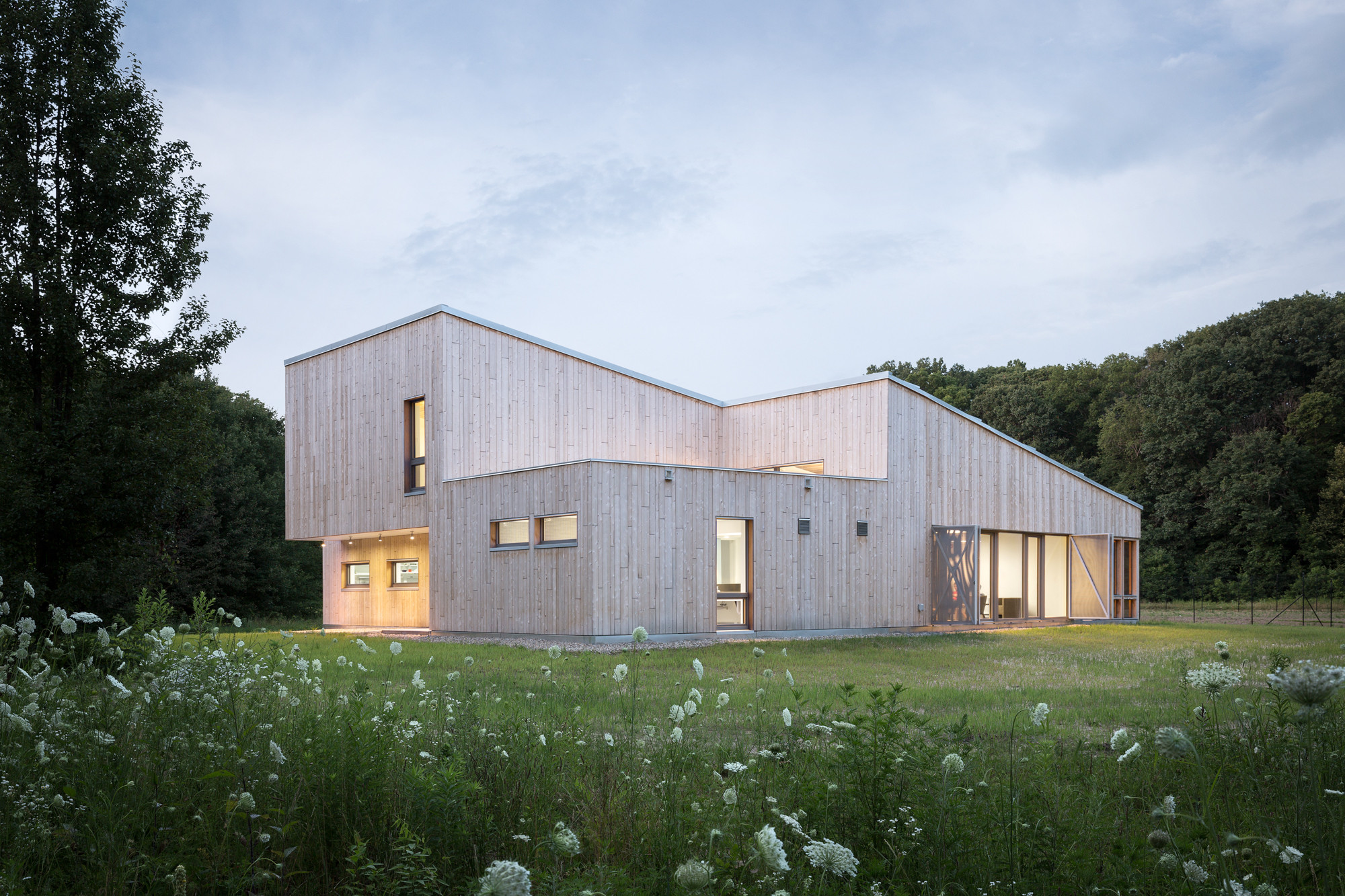 Das kleine, ein- bis zweigeschossige Gebäude steht am Rand einer großen Lichtung und hat eine Fassade aus hellem Zedernholz