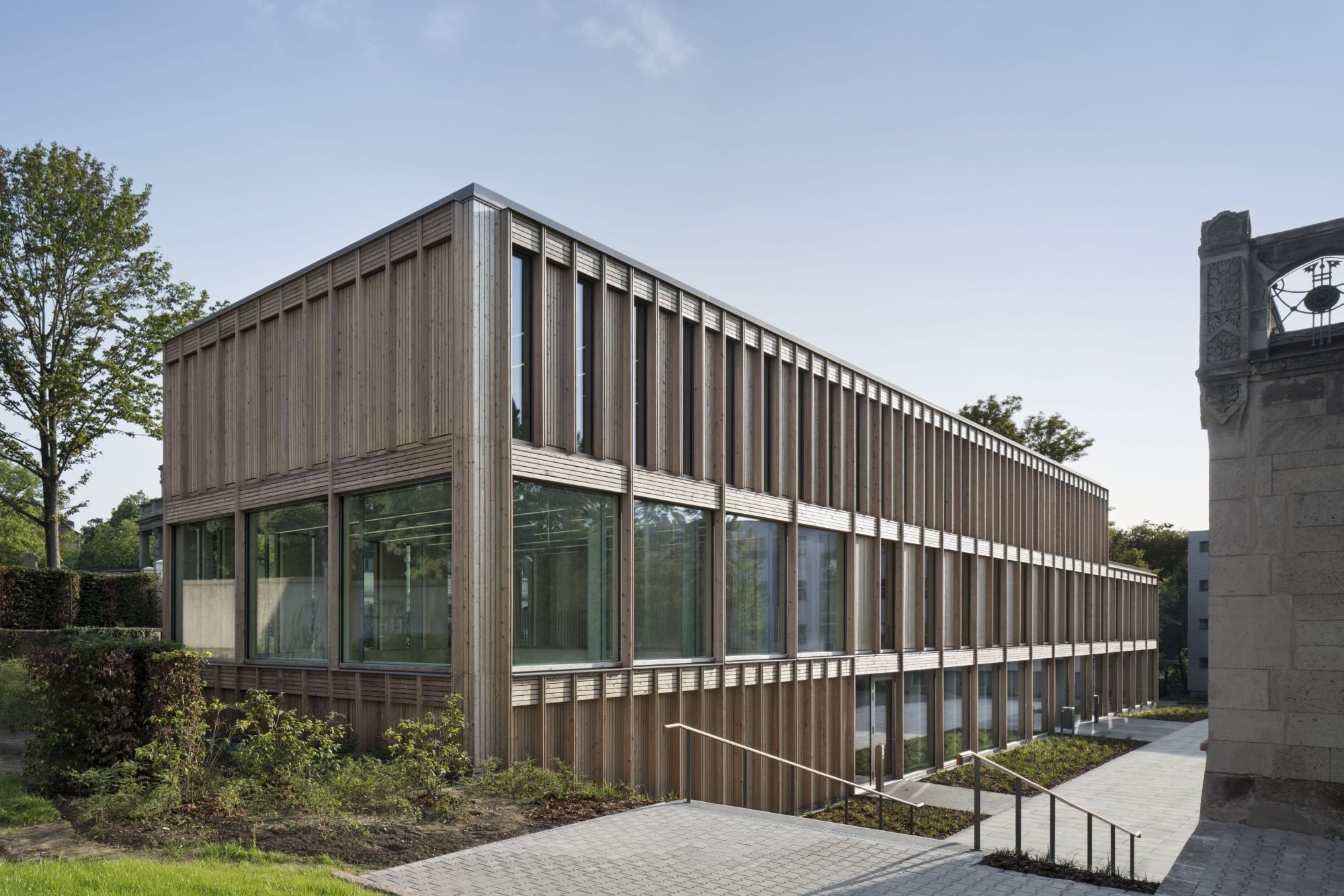 Zugang zum Campus: Der dreigeschossige Flachbau mit seiner rhythmisch gegliederten Holzfassade weckt Assoziationen an eine Remise oder Orangerie (Ansicht Nordost)