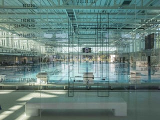 Die gläserne Schwimmhalle wird von zwei Trakten flankiert, die auch den Eingangsplatz im Süden fassen