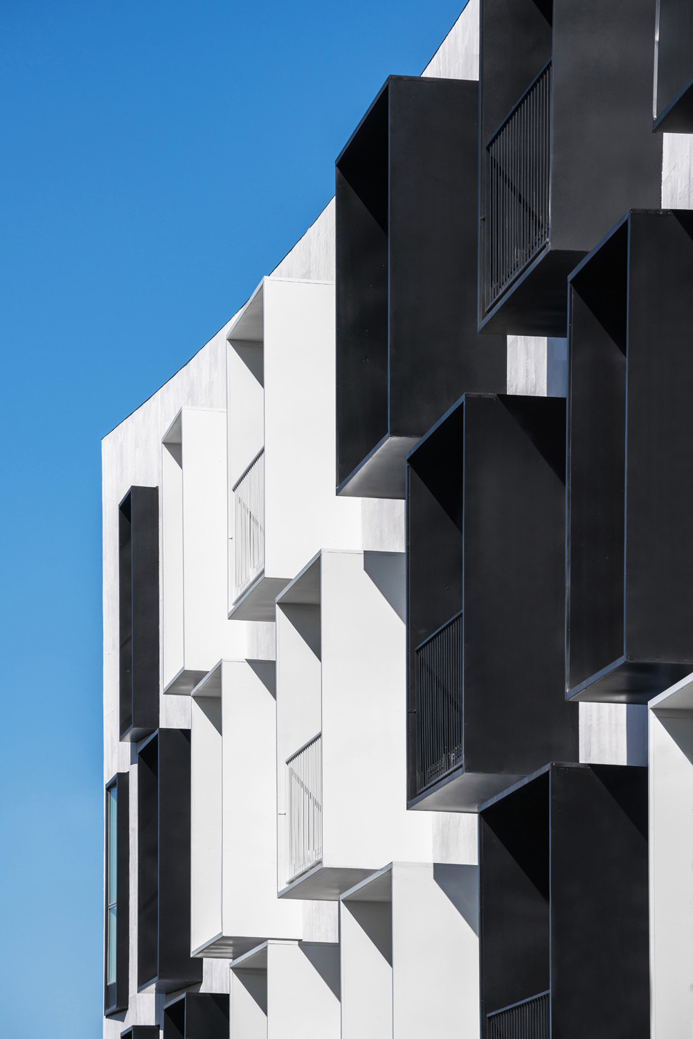 Die aufgesetzten Metallrahmen, die mal Loggien, mal schmale Austritte und auch mal kleine Fenster umfassen, ragen bis zur imaginierten geometrischen Kante des verzogenen Quaders hervor