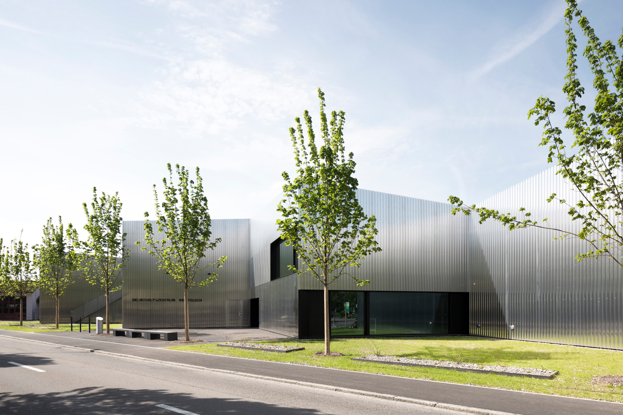 Das Gebäude auf dreieckigem Grundstück am Ortsrand reagiert zu den unterschiedlichen Seiten auf die angrenzenden Wohn- und Gewerbebauten. Zum Wohngebiet nach Nordosten ist das Volumen abgestaffelt und die Fassade mit Aluminiumtrapezblechen verkleidet