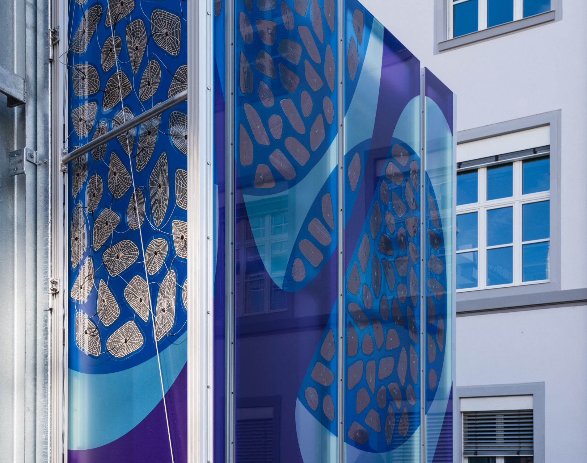 Teils transparent, teils blau und violett ist sie gemäß den CI-Vorgaben des Auftraggebers mit sich überschneidenden Blasenformen bedruckt, in die sich die ebenfalls organisch geformten OPV-Module, die an Spinnennetze denken lassen, wie selbstverständlich einfügen