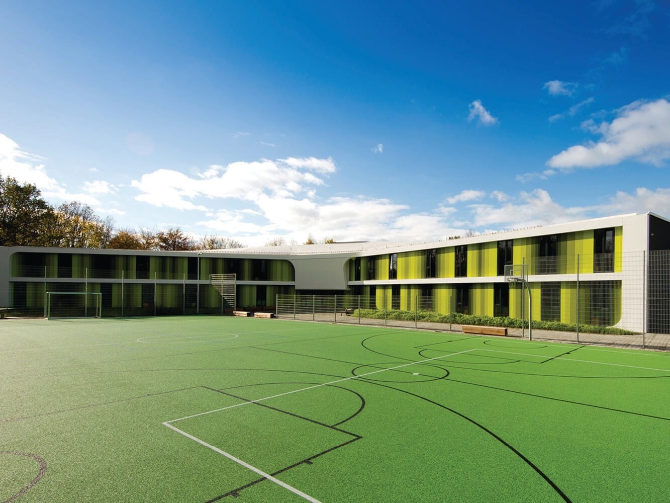 Wie ein Ypsilon ruht der Baukörper auf einem von Bäumen gesäumten Grundstück. Zwischen den Flügeln befinden sich je ein Sportfeld, ein Abenteuerspielplatz sowie Grün- und Verweilzonen.