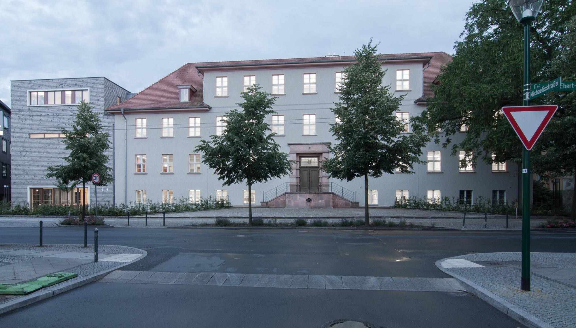 Der hölzerne Anbau schließt direkt östlich an das bestehende Hauptgebäude, ein dreigeschossiges Baudenkmal mit Putzfassade, an.