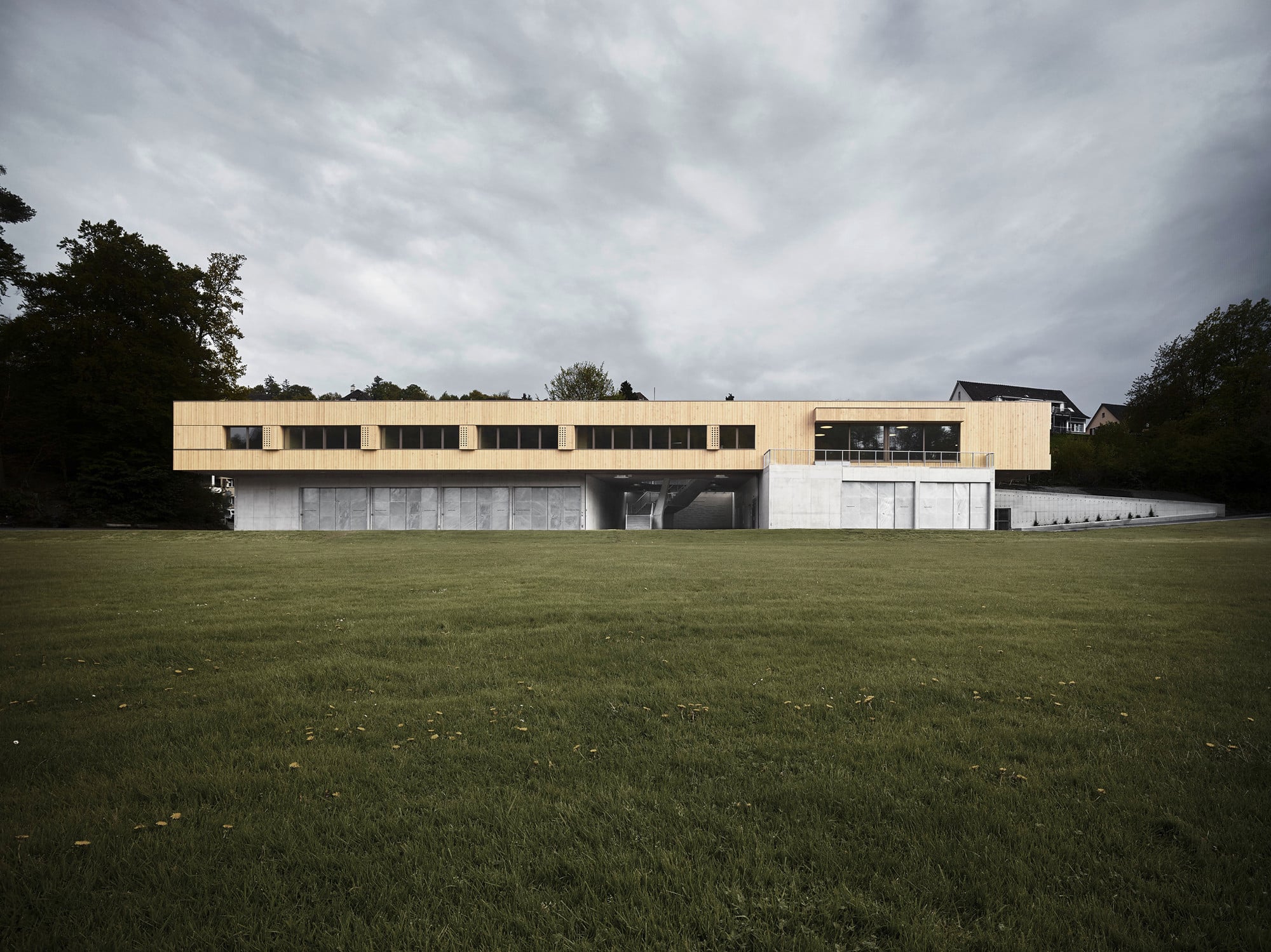 Das Gebäude besteht im Erdgeschoss aus zwei leicht schräg zueinander angeordneten Volumina aus Beton, auf denen eine leichte, langgestreckte Holzkonstruktion ruht.