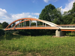 Bogenbrücke bei Lohmar Höngesberg / Kreuznaaf: Das Haupttragwerk – die beiden Bögen – besteht aus blockverleimten Brettschichtholz (BSH)-Balken (Fichte), die seitlich mit Lärchenholz verkleidet sind. Auch die beiden Vorlandbrücken sind aus blockverleimten Fichten-BSH gefertigt. Den statische Verbund zur oberseitigen Ortbetonplatte gewährleisten in das Holz eingeklebte Verbindungsmittel. Um das Bauwerk vor Witterungseinflüssen zu schützen, sind die Bögen oberseitig mit Titanzinkblechen abgedeckt und seitlich verkleidet. Exponierte Bauteile wie zum Beispiel die Hänger und das Geländer sind aus Stahl hergestellt. Der Handlauf besteht aus Accoya-BS-Holz.