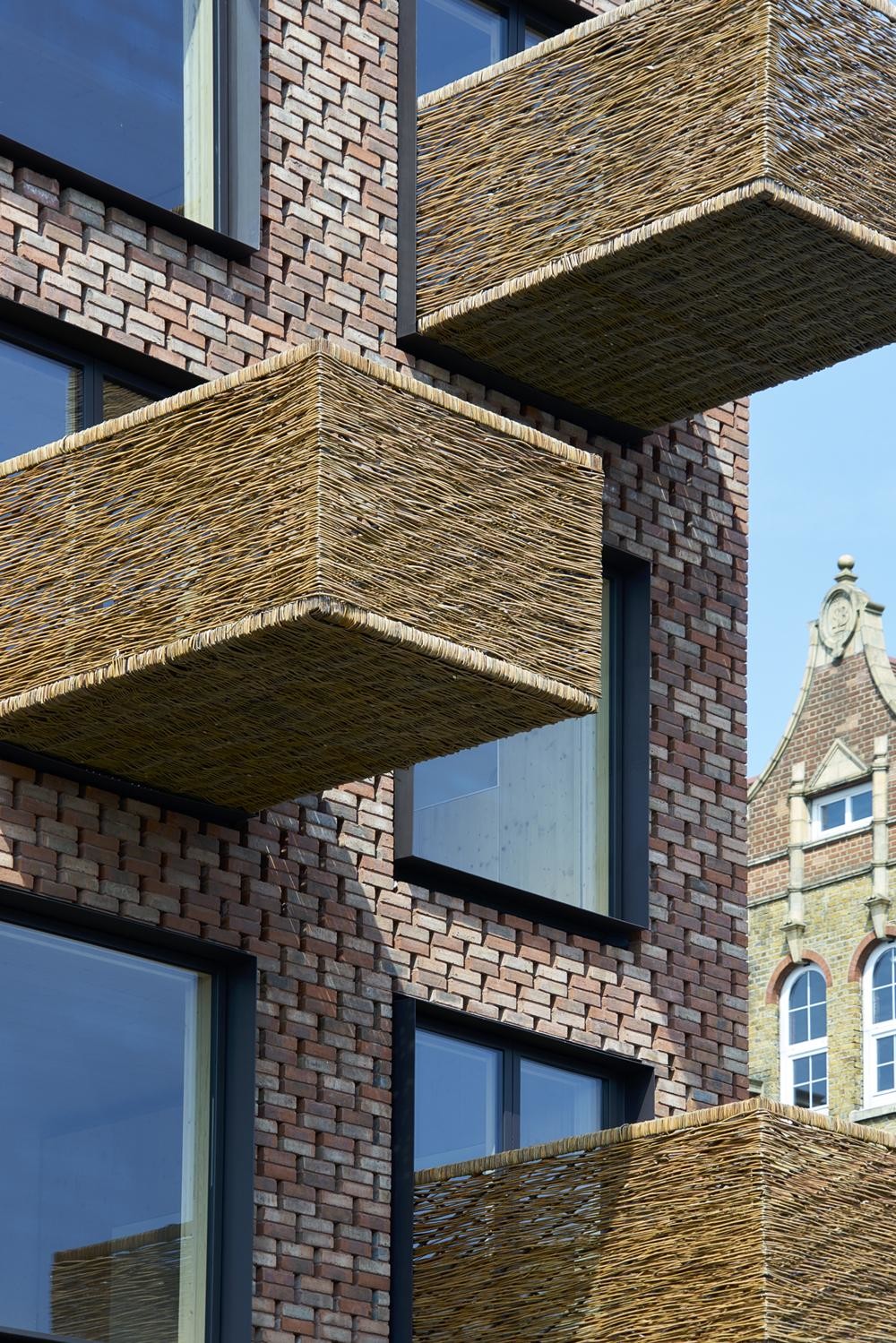 Ziegelsteine bekleiden als Vormauerschale sowohl die Fassade als auch das Satteldach. Die Balkonbrüstungen aus Weidengeflecht ragen markant ins Straßenbild.