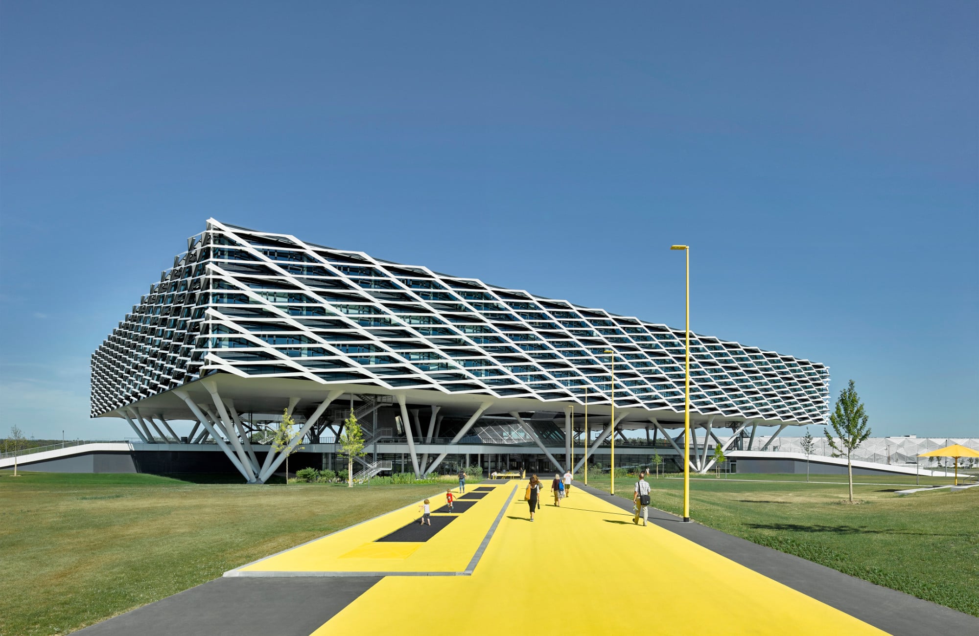 Bei dem dreigeschossigen Neubau handelt es sich um ein Empfangs- und Bürogebäude. Hinter den weißen Sonnenschutzlamellen verbirgt sich eine preisgekrönte Stahl-Beton-Konstruktion, die auf 67 Stahlstützen liegt.
