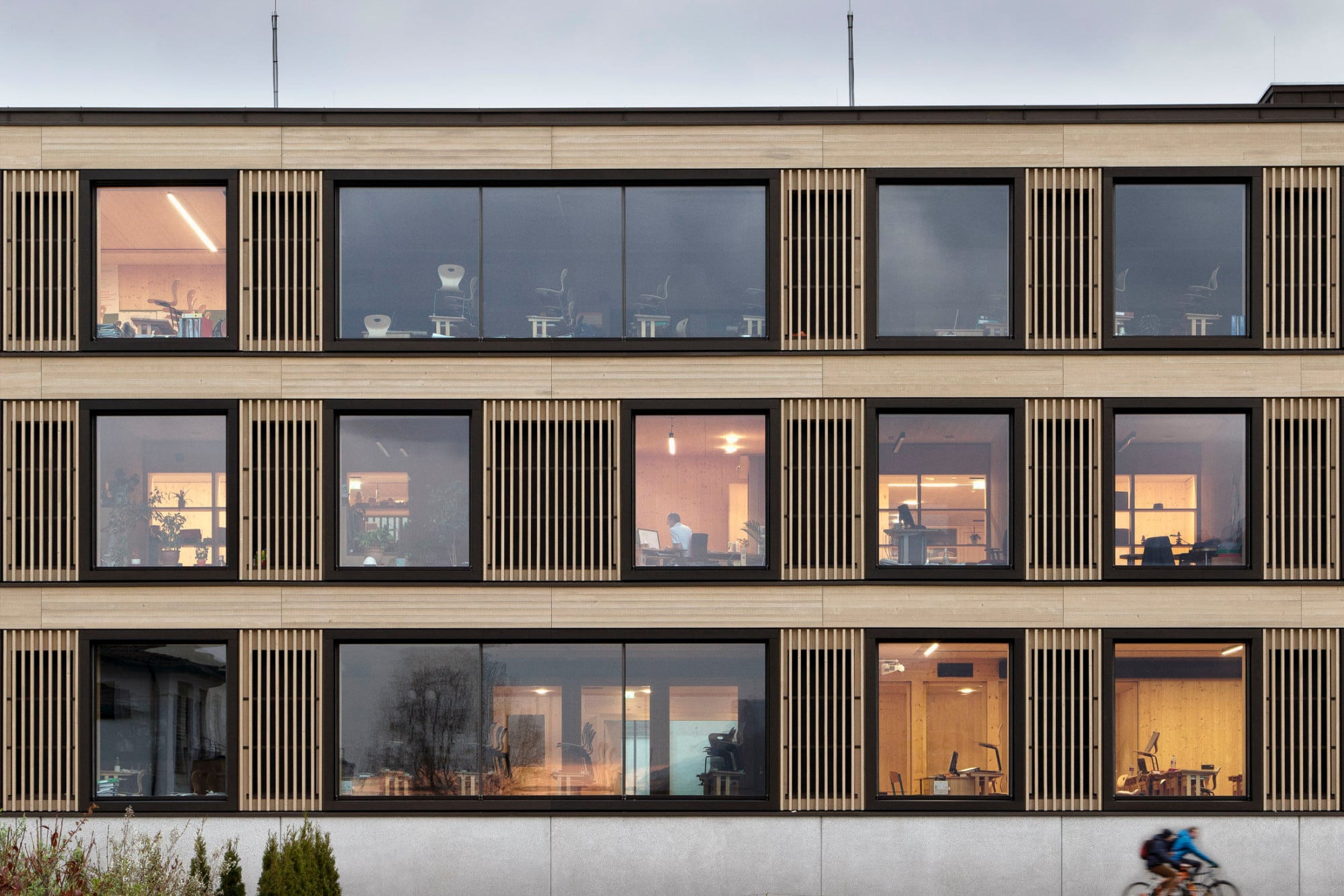 Der Sockel ist aus vorgefertigten Sichtbetonbauteilen errichtet, die Geschosse darüber aus Holz: Die Fassade ist geprägt durch große Fenster und vertikale Lamellen, hinter denen sich Öffnungsflügel verbergen. 