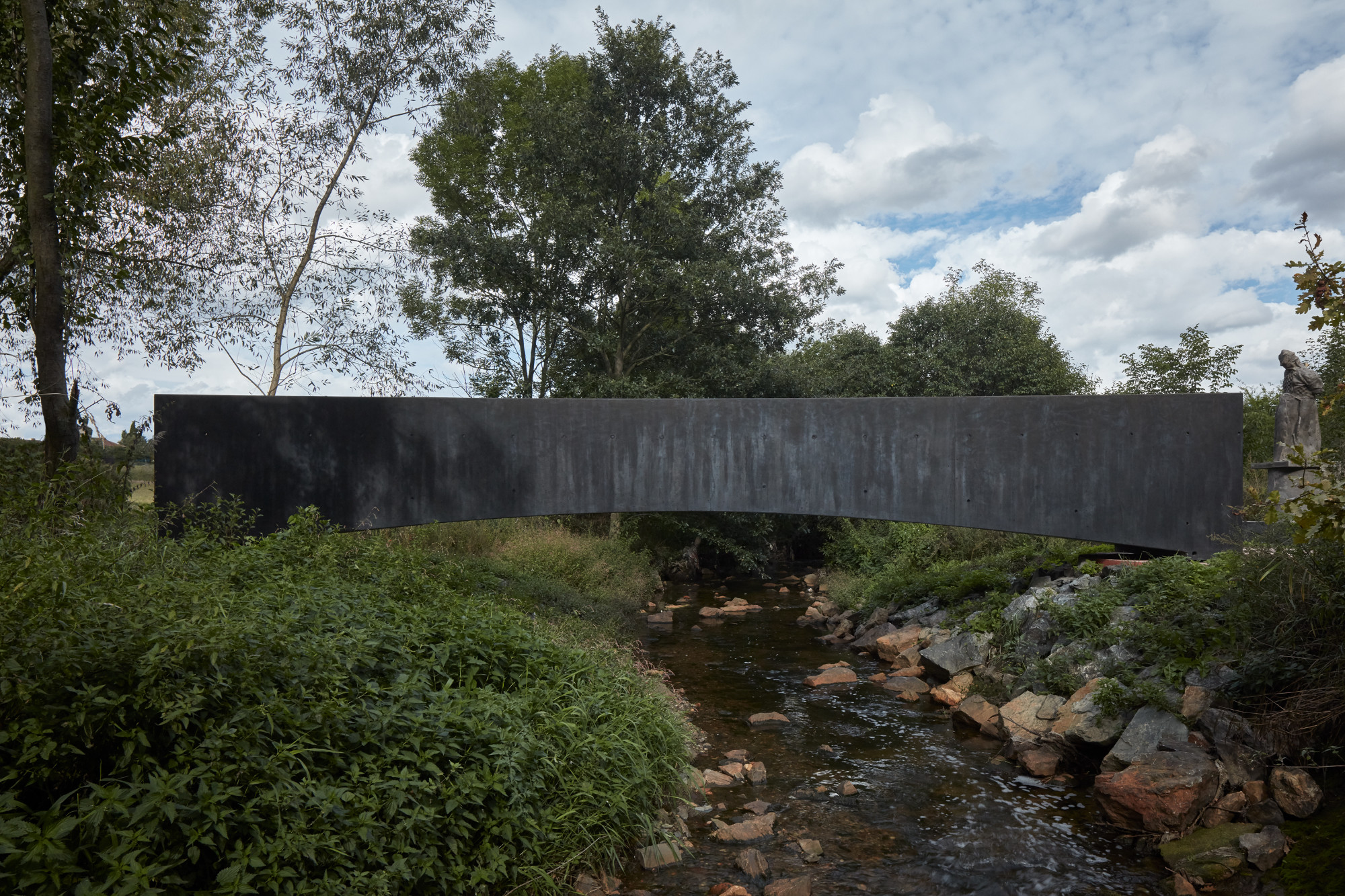 Die Brücke spannt in einem sanften Bogen über das naturbelassene Gewässer, das regelmäßig über die Ufer tritt und die angrenzenden Wiesen überschwemmt.