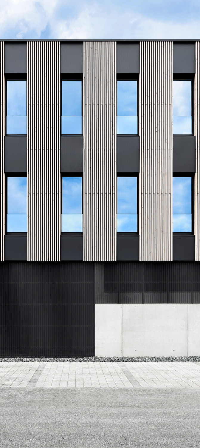 Die strenge Geometrie des Baus wird durch die Gestaltung der Gebäudehülle zusätzlich unterstützt: Vertikale Holzlamellen geben der homogenen Fassade eine plastische Struktur. 