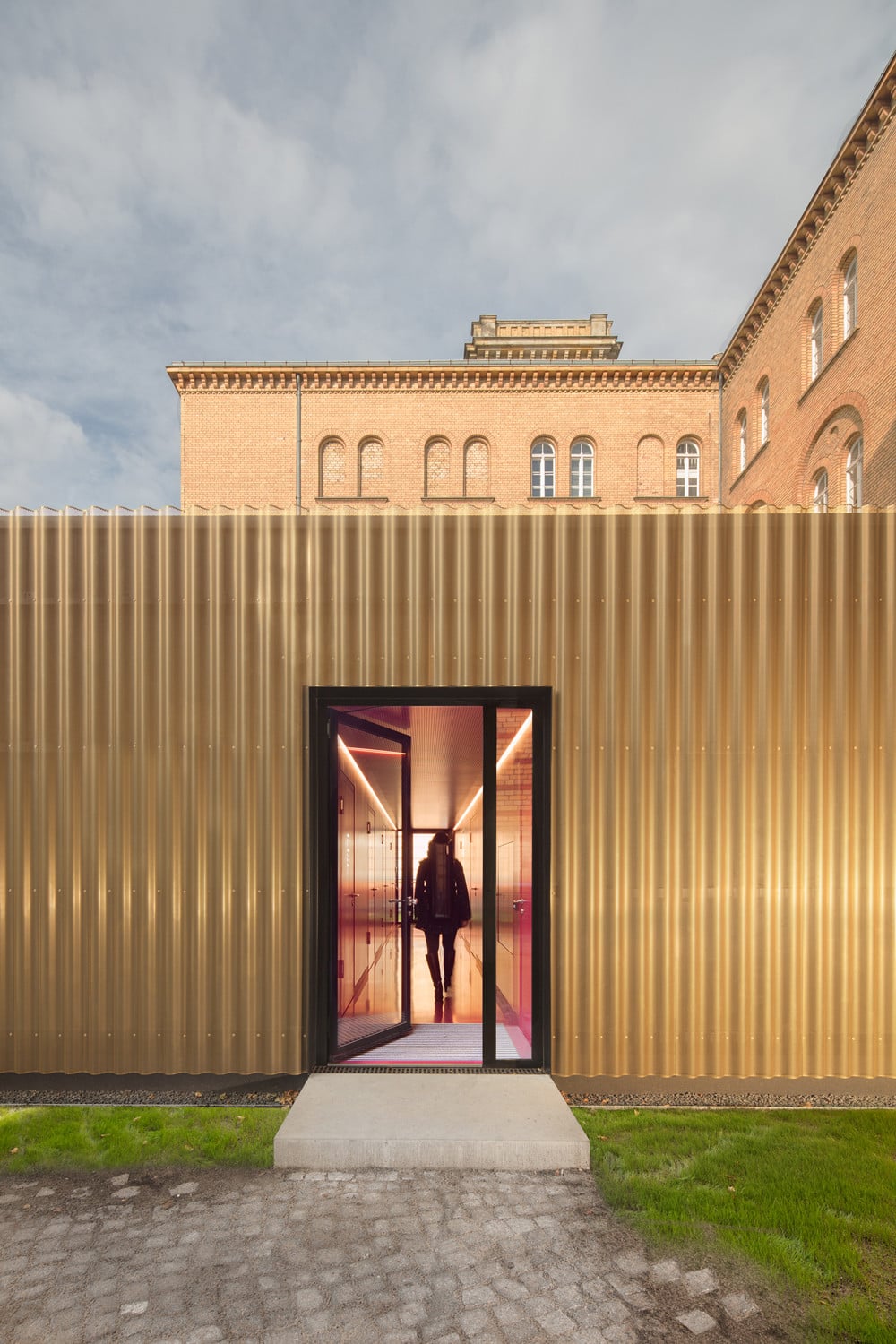 Die golfarbene Fassaden aus perforiertem, eloxiertem Wellblech hüllen die vorgefertigten Holzmodule ein und greifen den warmen Farbton der umliegenden Klinkerwände auf.