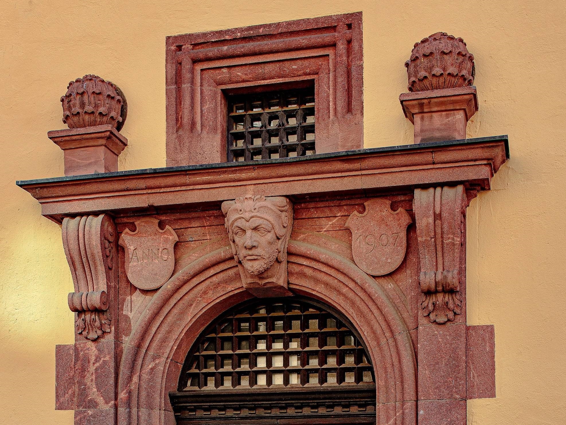 Kartusche mit Segensspruch, Zunftzeichen und stilisiertem Löwenkopf, unten Ranken aus Eichenlaub und Akanthus. Vermutlich hatten auch die nun leeren Flächen auf den Säulenköpfen ursprünglich Reliefs mit symbolischer Darstellung.