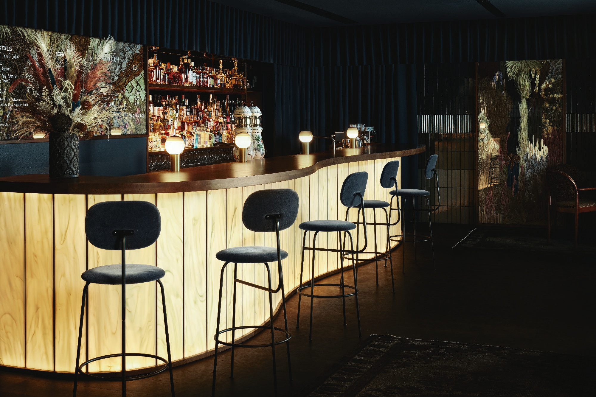 Beim Betreten der Bar fallen zunächst das schummrig gedimmte Licht und die dunkle Farbpalette auf, aus der die geschwungene, hinterleuchtete Tresenfront im Zentrum des Raumes als strahlender Fixstern hervorsticht. 