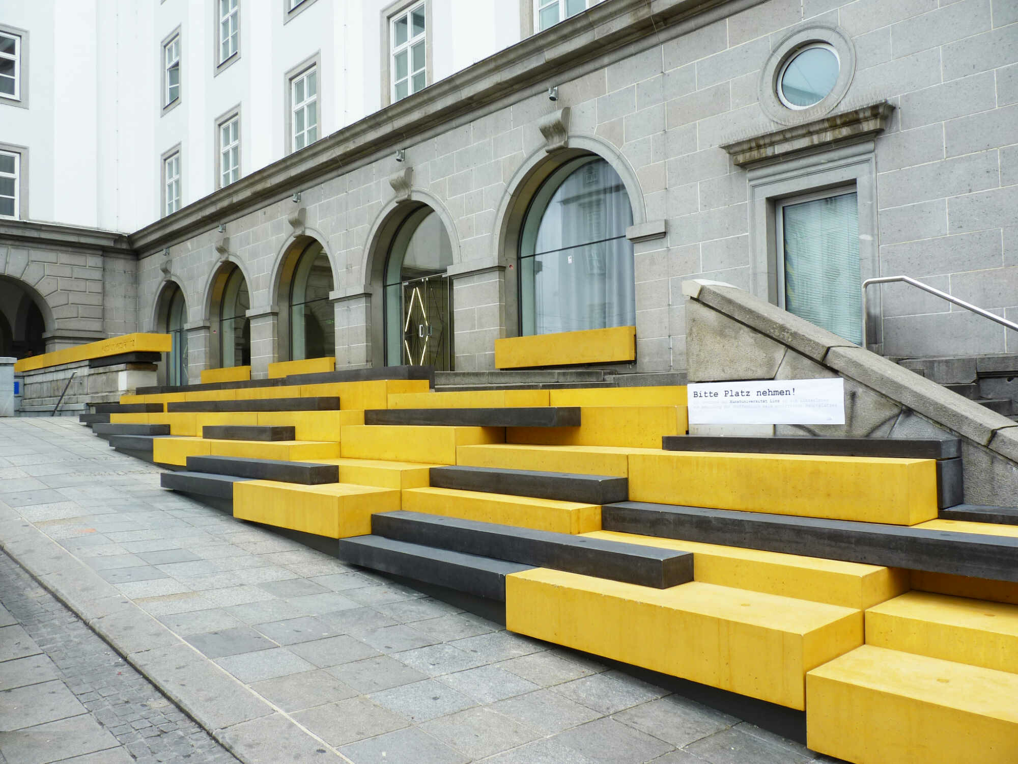 Sitzstufen in Schwarz und Gelb laden vor dem Haupteingang der Linzer Kunstuniversität zum Verweilen ein.
