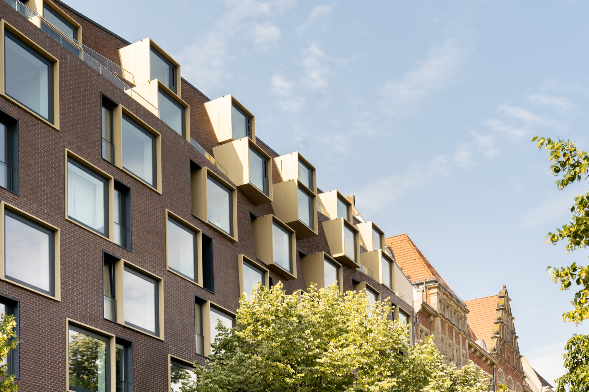 Die streng quadratischen Fenster, deren messingfarbene, metallene Rahmen an einigen Stellen erkerartig auskragen, durchbrechen die organische Fassade.