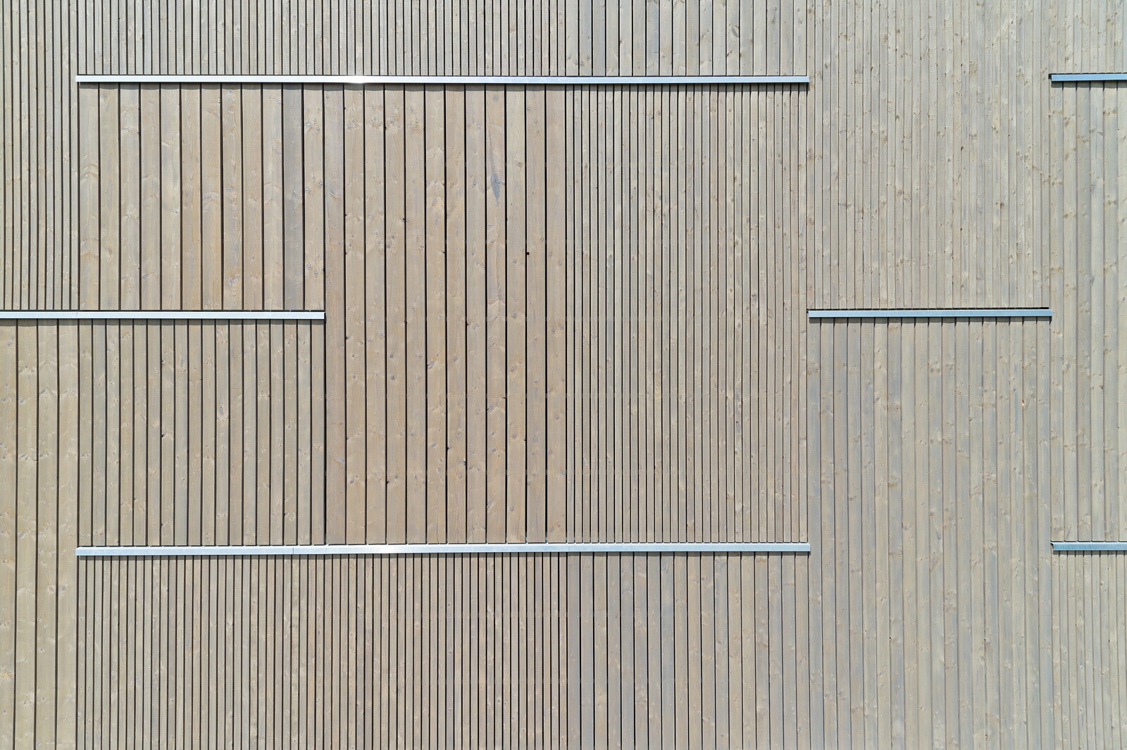 Nähert man sich jedoch dem Gebäude, wird die dezente Rhythmisierung der Flächen durch die unterschiedlich breiten Bretter erkennbar, die zudem durchbrochen wird von horizontalen Aluminiumleisten.
