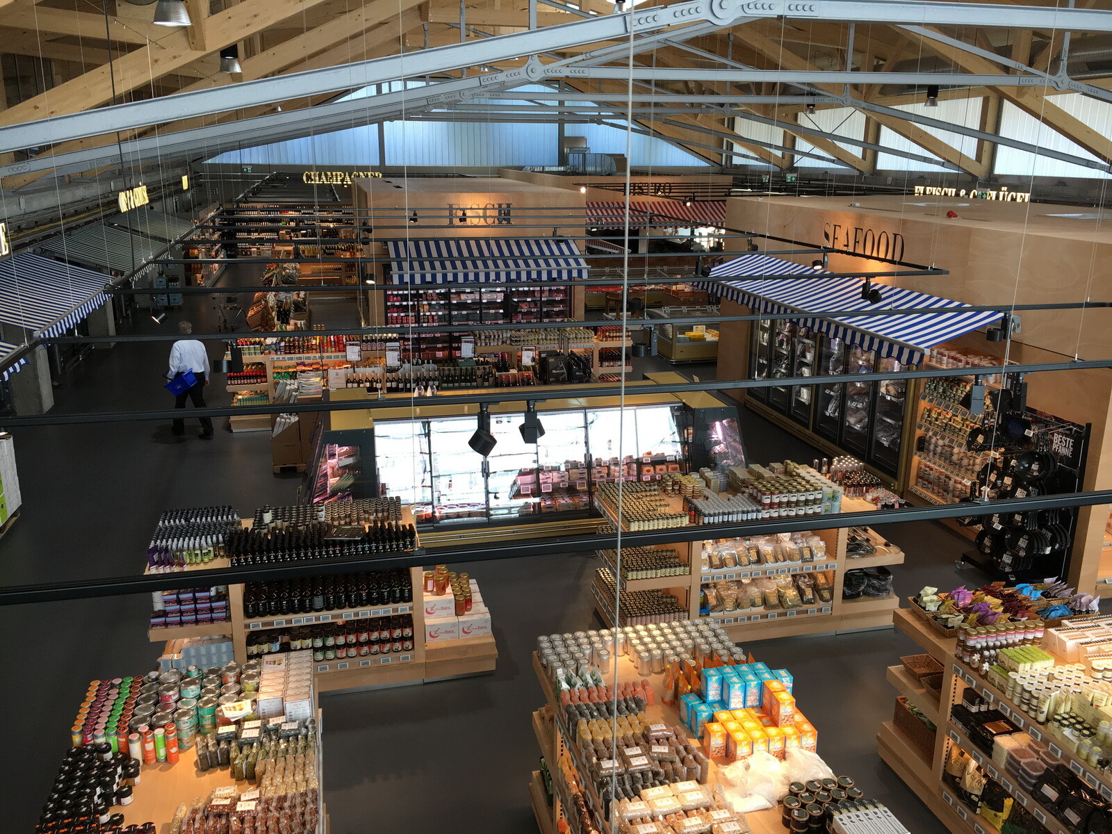 Der Innenraum ist geprägt von den unbehandelten Rohbau-Holzbindern, dem dunkelgrauen Industrieboden und den hölzernen Regalen und eingestellten Ständen, die an eine Markthalle denken lassen. 