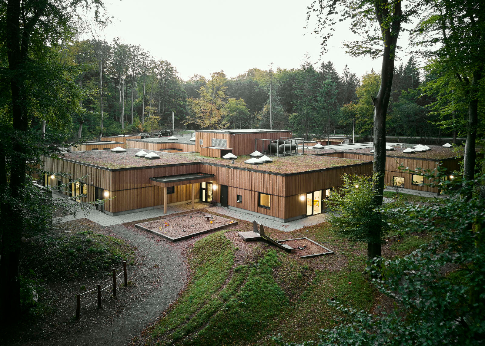 Ein hügeliger Spielplatz mit alten Bäumen umgibt das Gebäude. Möglichst viel Baumbestand wurde belassen und die notwendigerweise für den Baugrund gefällten Bäume wurden als Gestaltungselemente auf dem Spielplatz wiederverwendet.