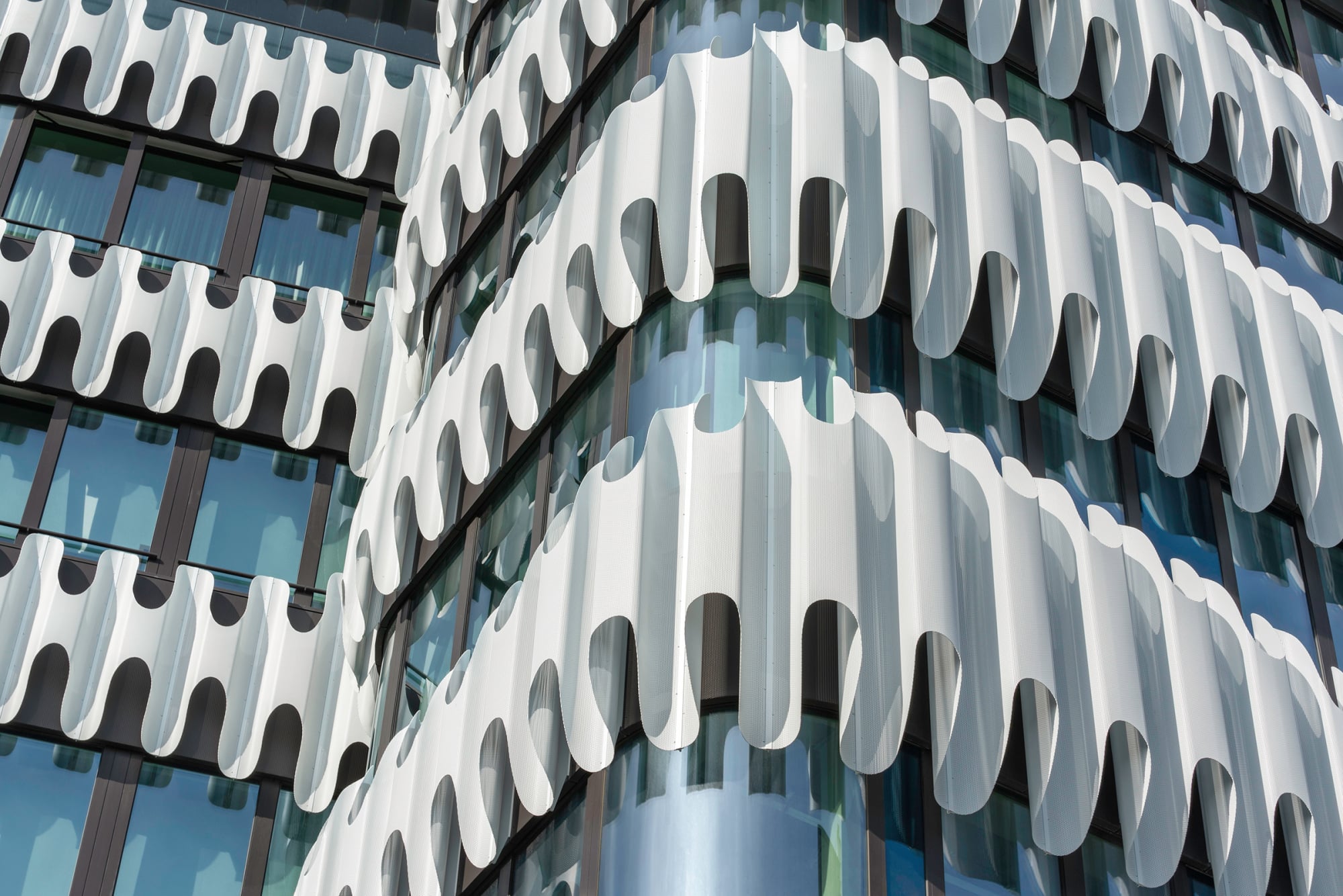 Als Betonkonstruktion errichtet, ist das Hochhaus umhüllt von einer vollflächigen Glasfassade. Diese wiederum ist umgeben von umlaufenden weißen Wellenbändern aus Aluminium.