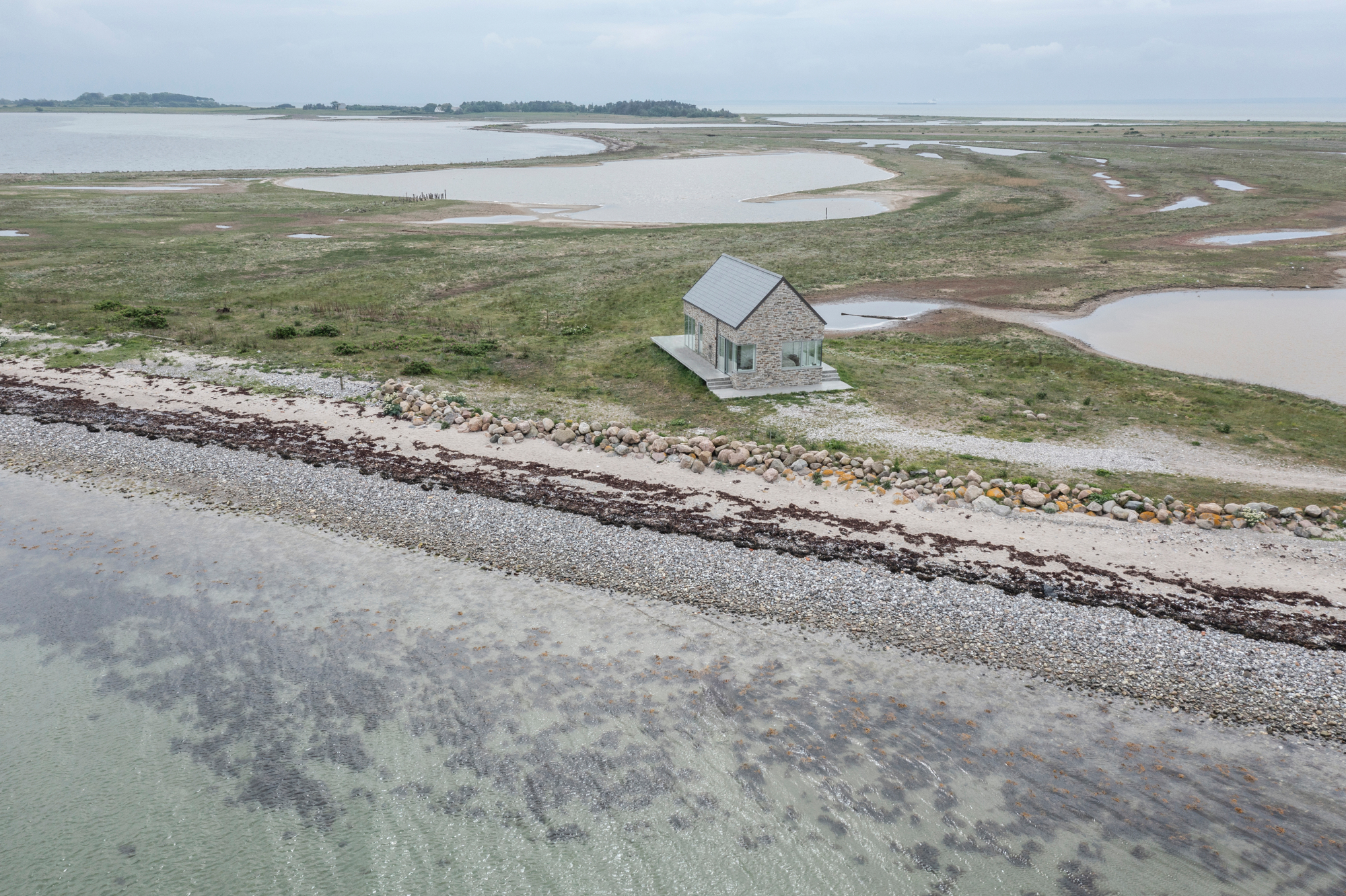Nur ein schmaler Küstenstreifen mit Sand, Kieseln und größeren Steinen liegt zwischen dem Meer und dem schlicht wirkenden Sommerhaus. 
