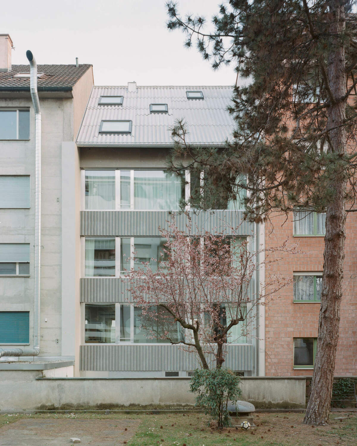 Horizontale Bänder aus anthrazitfarbenem gewelltem Faserzement und große außenbündige Aluminiumfenster gliedern die minimalistische Fassade.