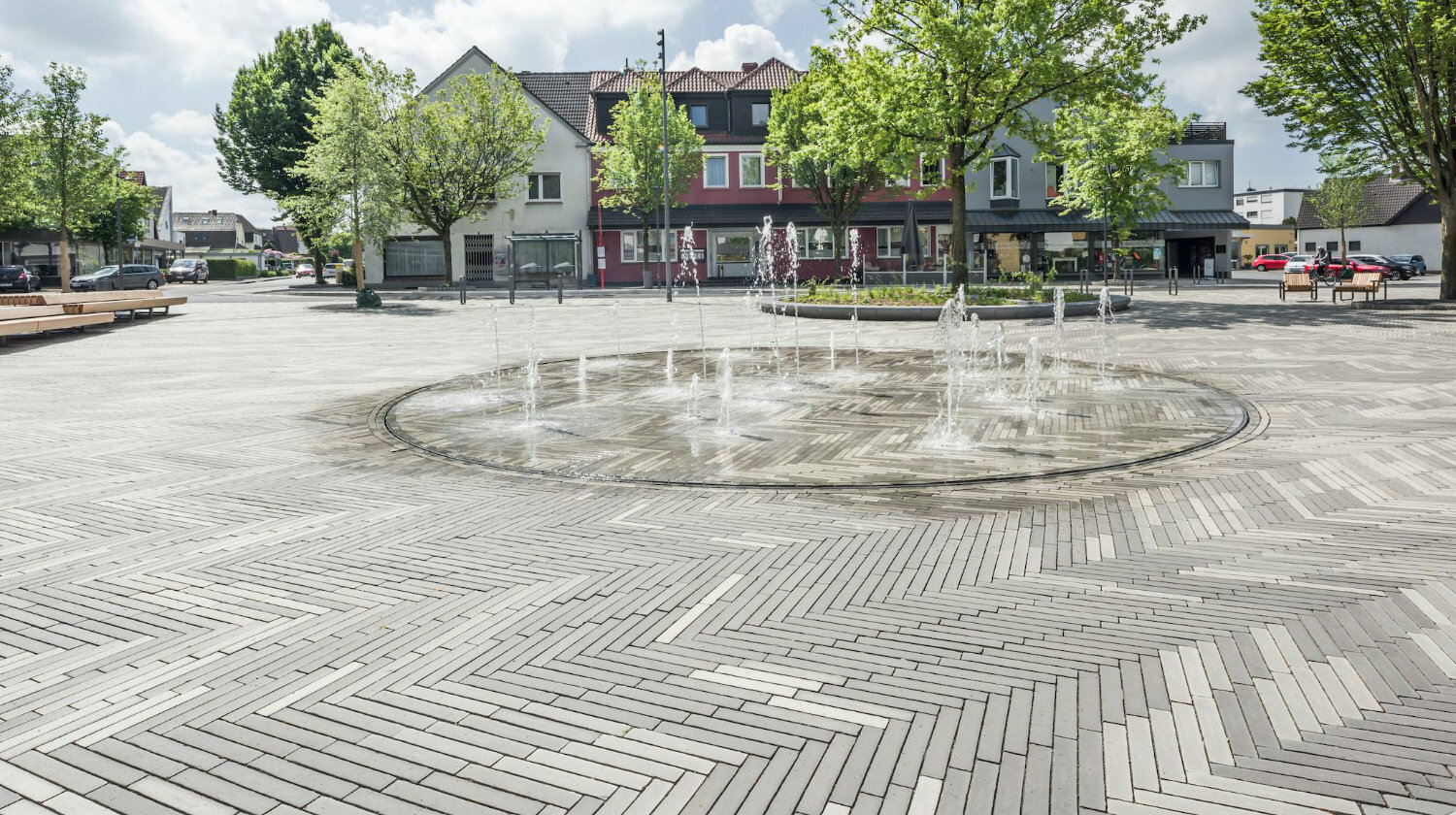 Ein Wasserspiel bildet die Mitte des Platzes, der von Bäumen umgeben ist.