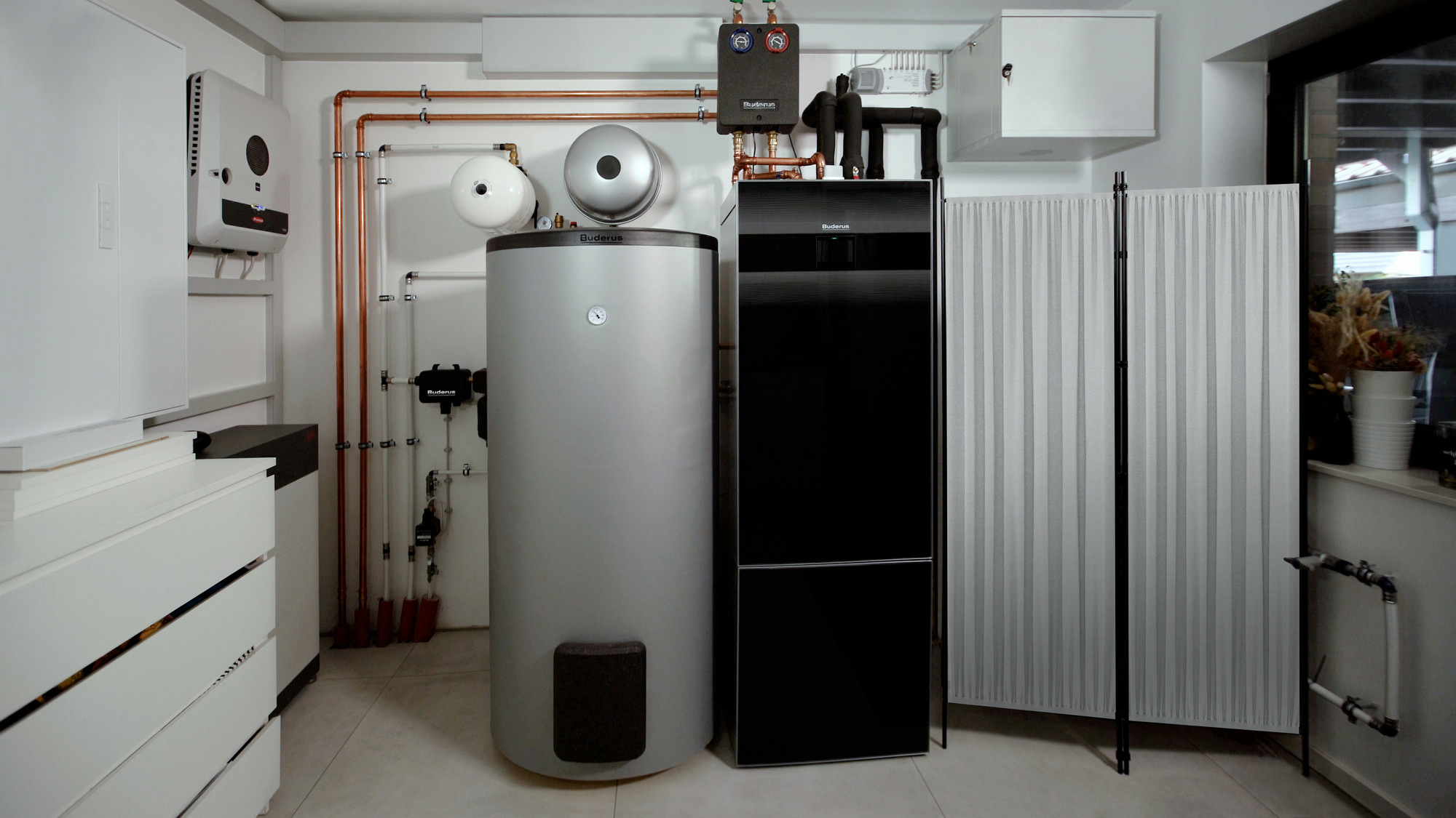 Zentrale Komponente der Heizwärme- und Warmwasserbereitung ist die Sole-Wasser-Wärmepumpe Logatherm WSW196i (Mitte). Links daneben stehen ein Pufferspeicher sowie ein Batteriespeiche an der Wand.