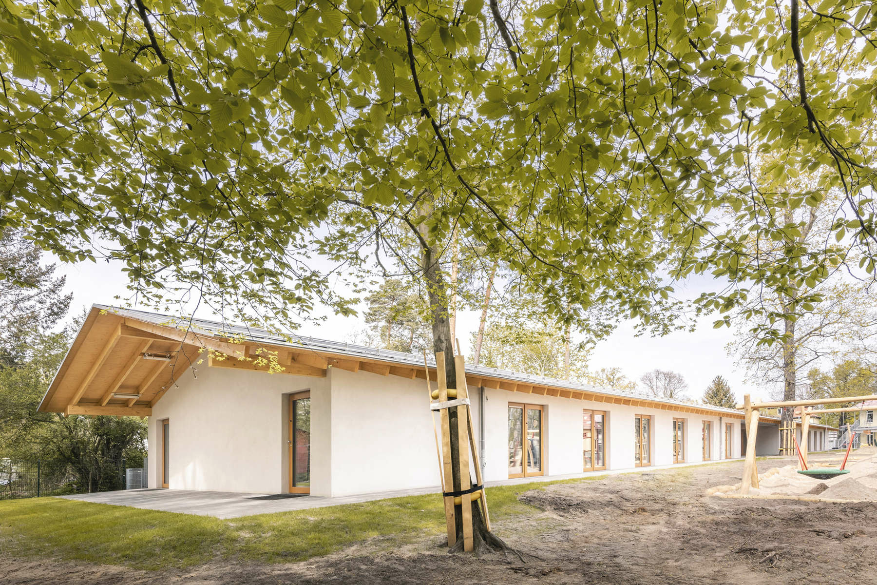 Mit einer natürlichen Material- und Farbwahl und dem flach geneigtem Satteldach mit großen Überständen fügt sich der lange, eingeschossige Neubau behutsam in die dörfliche Idylle ein. 