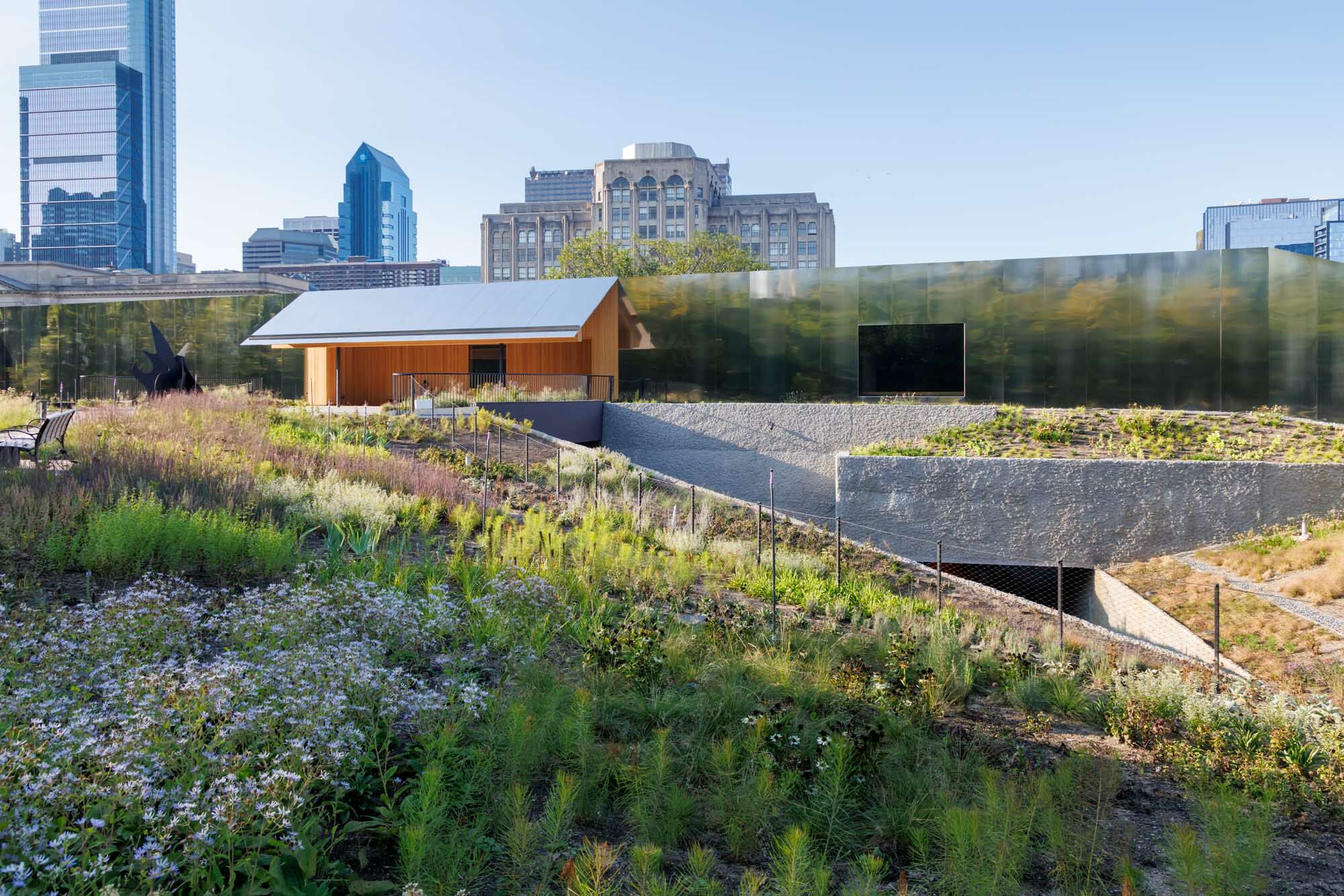 Ein Weg führt durch eine wiesenartige Landschaft auf den mit Metallpaneelen verkleideten Museumsbau zu, in dessen Fassade sich die aufwendig gestaltete Gartenlandschaft spiegelt.