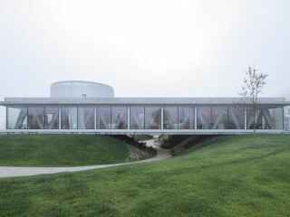 MVA Mikelic Vres Architekten planten einen geradlinigen eingeschossigen Baukörper, der die organisch geschwungene Topografie überlagert.. 