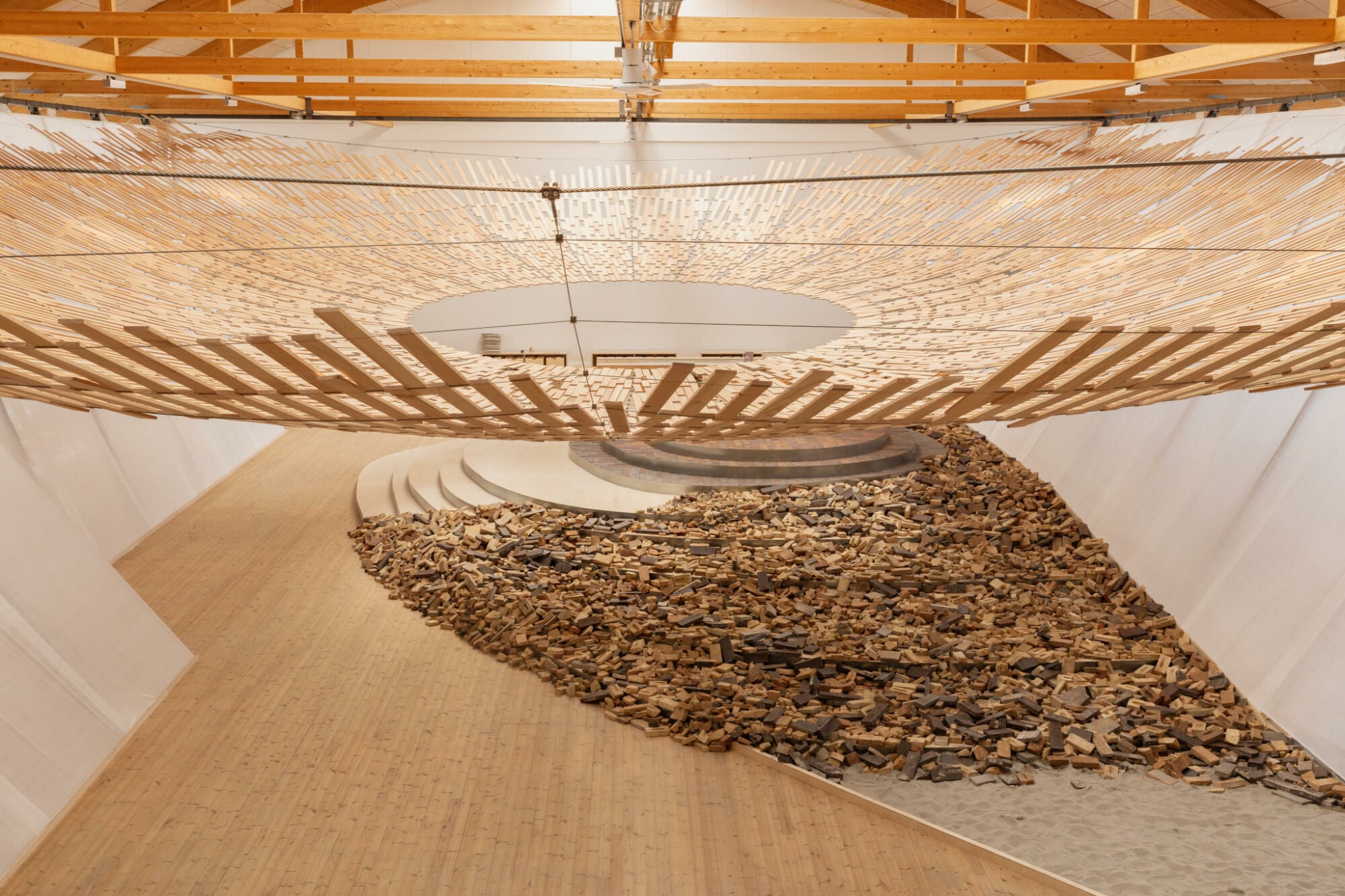 Zentrales Element der Ausstellung ist eine schalenförmige, konvexe Deckenkonstruktion aus wiederverwendeten, lückenhaft gefügten Holzleisten.