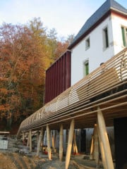 Als eigenständiger Teil bildet die neue Holzterrasse den Übergang zwischen dem Muralen und der Natur. Skulptural anmutend drängt sie nach vorne: zur Aussicht an den Rand des steilen Geländes.