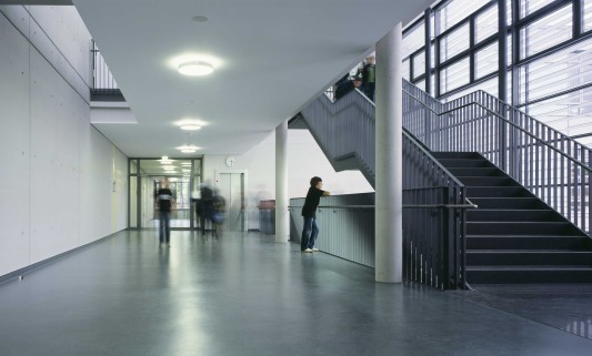 Große Fensterfronten, weiße Wänden und Säulen aus Sichtbeton stehen in den Fluren in Kontrast zum schwarzen Linoleumboden